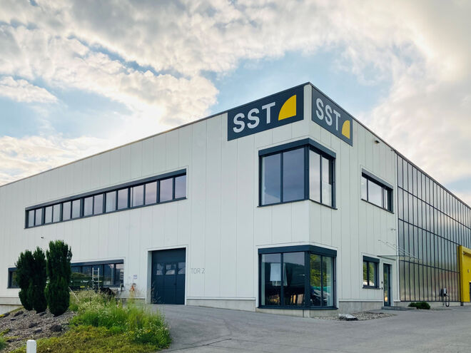 SST Betriebsgebäude Ein modernes Industriegebäude mit der Aufschrift "SST" auf der Fassade, umgeben von einer gepflasterten Fläche und einigen Pflanzen, unter einem bewölkten Himmel. SST Betriebsgebäude Ein modernes Industriegebäude mit der Aufschrift "SST" auf der Fassade, umgeben von einer gepflasterten Fläche und einigen Pflanzen, unter einem bewölkten Himmel.