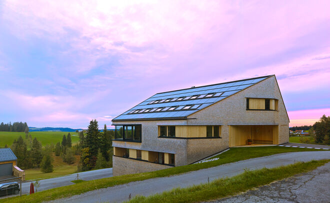Modernes Haus mit geneigtem Dach und Solarpanelen, umgeben von grünen Wiesen und Bäumen. Der Himmel zeigt ein beeindruckendes rosa-blaues Farbspiel. Modernes Haus mit geneigtem Dach und Solarpanelen, umgeben von grünen Wiesen und Bäumen. Der Himmel zeigt ein beeindruckendes rosa-blaues Farbspiel.
